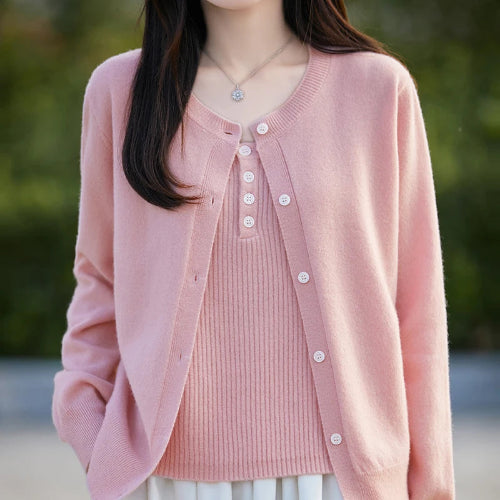 A woman wearing a light pink open-front cardigan over a matching pink ribbed top, photographed outdoors with a softly blurred green background.