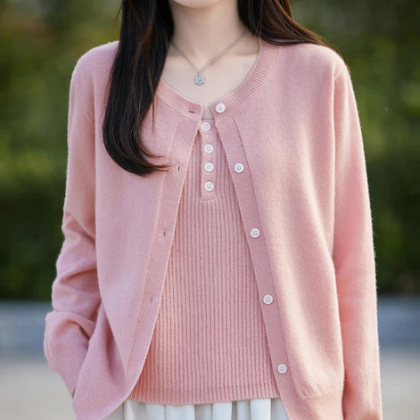 A woman wearing a light pink open-front cardigan over a matching pink ribbed top, photographed outdoors with a softly blurred green background.