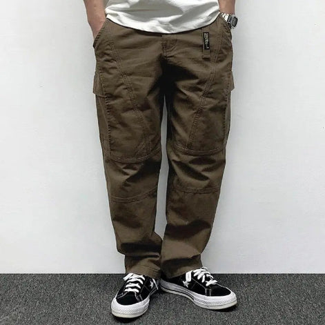 Front view of a model wearing brown loose-fit cargo pants with thigh pockets, curved seam detailing, and stitched knee panels, paired with black sneakers.⠋