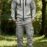 A man wearing a light gray zip-up hoodie and matching gray jogger cargo pants with white sneakers, standing in a sunlit forest clearing.