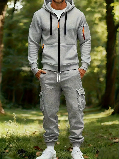 A man wearing a light gray zip-up hoodie and matching gray jogger cargo pants with white sneakers, standing in a sunlit forest clearing.