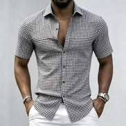 A man wearing a black and white gingham short-sleeve button-up shirt with white pants, standing against a light gray background.