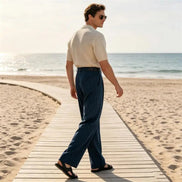 A man wearing a cream short-sleeve button-up shirt and dark blue pinstriped trousers walks along a wooden boardwalk on a sandy beach.