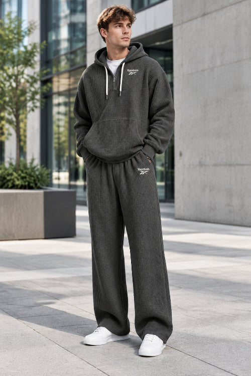 A man wearing a dark gray waffle-knit hoodie and matching sweatpants set with white Reebok logos, shown against a plain white background.