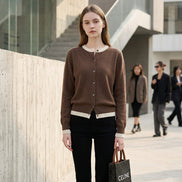A person taking a mirror selfie in an indoor room, wearing a brown button-front cardigan over a white top and black pants, while holding a black Celine shopping bag.