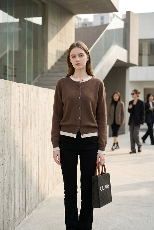 A person taking a mirror selfie in an indoor room, wearing a brown button-front cardigan over a white top and black pants, while holding a black Celine shopping bag.