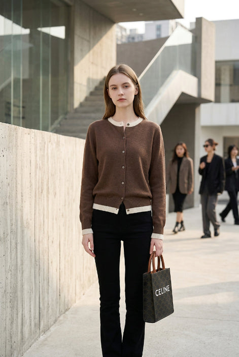 A person taking a mirror selfie in an indoor room, wearing a brown button-front cardigan over a white top and black pants, while holding a black Celine shopping bag.