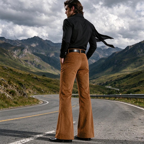 A man stands on a winding mountain road wearing a black long-sleeve shirt and brown flared pants, photographed from behind with mountains and a cloudy sky in the background.