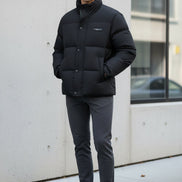 A black puffer jacket hanging on a clothing rack in a retail store setting, with promotional graphic overlays on the left highlighting warmth, softness, and down content. The jacket has a high collar, front zipper and snap closure, and a small chest label.
