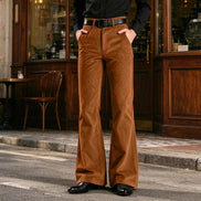 A person wearing brown wide-leg corduroy pants with a black belt, paired with a black long-sleeve shirt and black shoes, standing on a street in front of a wooden storefront.