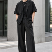 A two-piece men's casual outfit set in black, featuring a short-sleeve blazer-style jacket with rolled sleeves and a matching pair of wide-leg trousers, 