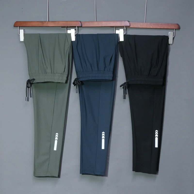 Three pairs of pants in green, blue, and black hanging on a rack against a gray background.