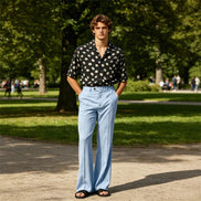 A man stands in a sunny park wearing a black short-sleeve button-up shirt with white polka dots, light blue wide-leg trousers, and black sandals.