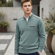 A man wearing a sage green quarter-zip knit sweater with shoulder detailing, styled over a white collared shirt and navy trousers, standing in a modern outdoor walkway.