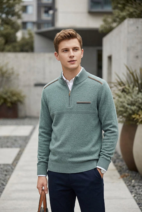 A man wearing a sage green quarter-zip knit sweater with shoulder detailing, styled over a white collared shirt and navy trousers, standing in a modern outdoor walkway.