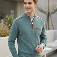 A man wearing a teal-green ribbed quarter-zip sweater with brown shoulder and chest accents, styled over a white collared shirt and black trousers, standing in a modern outdoor patio setting.