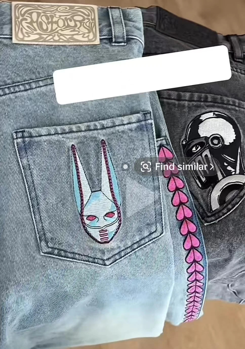 A close-up of a light-wash denim pair of jeans laid flat on a wood surface, showing the back pocket with a stylized embroidered face patch, a pink heart-stitched border along the side seam, and another dark denim pair partially visible to the right with a black skull patch.