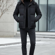 A man wearing a black puffer jacket with a brown-lined hood over a black turtleneck, shown against a dark gray background with feature callouts on the left.