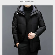 A man wearing a black hooded puffer jacket over a black turtleneck sweater, standing against a dark gray studio background. 