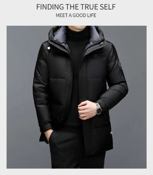 A man wearing a black hooded puffer jacket over a black turtleneck sweater, standing against a dark gray studio background. 