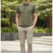 A close-up of a short-sleeve polo shirt in a muted olive-khaki color, hanging on a black hanger with a hand holding the fabric to show texture. The shirt features a classic collar, a three-button placket, and a small embroidered logo on the chest.
