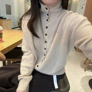 A person wearing a light beige ribbed turtleneck cardigan sweater with black buttons, photographed in a cafe setting.
