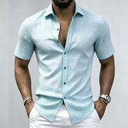 A man wearing a light blue and white gingham short-sleeve button-up shirt with white pants, shown from the chin down against a plain light gray background.