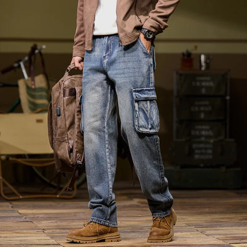 Front-angle view of a model wearing faded blue denim cargo pants with a large flap thigh pocket and straight legs, holding a brown backpack and wearing brown boots.