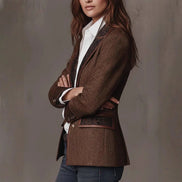 Side view of a woman with folded arms wearing a brown herringbone-pattern jacket with contrast lapels, pocket trim and buttoned cuffs over a white shirt