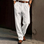 Person wearing white pleated pants and brown shoes against a textured wall.
