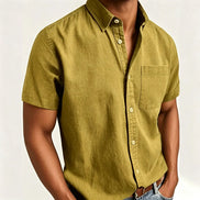 A man wearing a short-sleeve button-up shirt in a muted olive yellow-green color, paired with blue jeans and a brown leather belt,