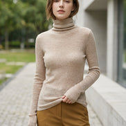 A woman wearing a beige ribbed long-sleeve turtleneck sweater, taking a mirror selfie in an indoor setting with a tiled wall in the background.