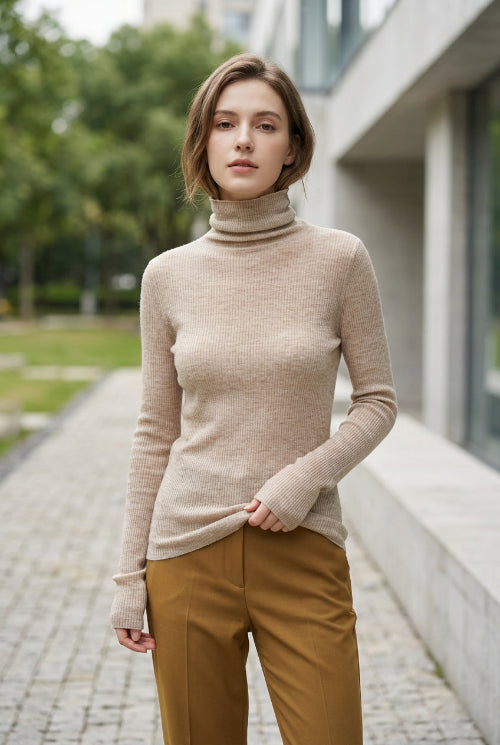 A woman wearing a beige ribbed long-sleeve turtleneck sweater, taking a mirror selfie in an indoor setting with a tiled wall in the background.