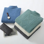 Three folded cable-knit mock-neck sweaters in blue, mint green, and heather gray, styled with a black leather wallet, a watch, and a magazine on a white background.