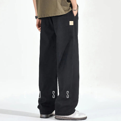 Rear view of a model wearing black loose-fit straight-leg pants with back patch pockets and a small sewn-on label, paired with white sneakers.