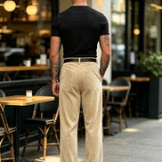 A man viewed from behind wearing beige linen trousers, a black t-shirt, and a black belt, standing on a patio in front of a cafe.