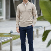 A man wearing a beige quarter-zip sweater, navy blue trousers, and white sneakers standing outdoors on a paved walkway.