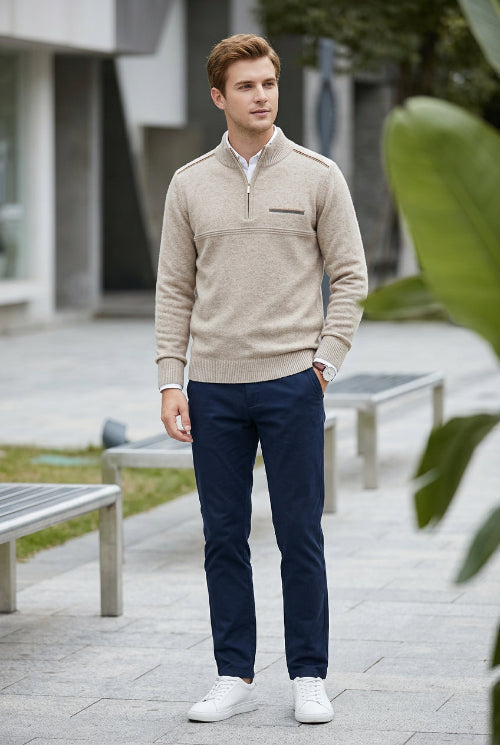 A man wearing a beige quarter-zip sweater, navy blue trousers, and white sneakers standing outdoors on a paved walkway.