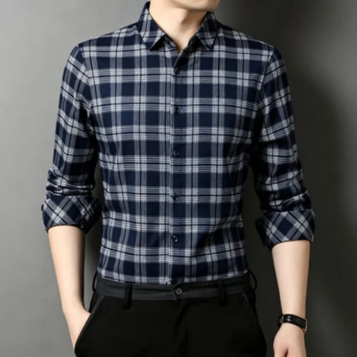 A man wearing a black and white plaid long-sleeve button-up shirt, paired with black trousers and a black belt, standing against a dark gray background.