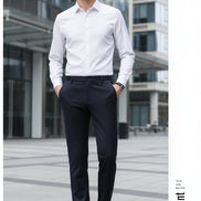 A man wearing slim-fit dark charcoal gray dress pants, styled with a white long-sleeve shirt and black leather dress shoes, standing against a dark gray studio background.