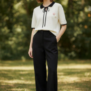 An off-white short-sleeve polo shirt with a black collar, black tie detail at the neck, and black piping on the sleeve cuffs. It features a small pink and white floral embroidery on the left chest.