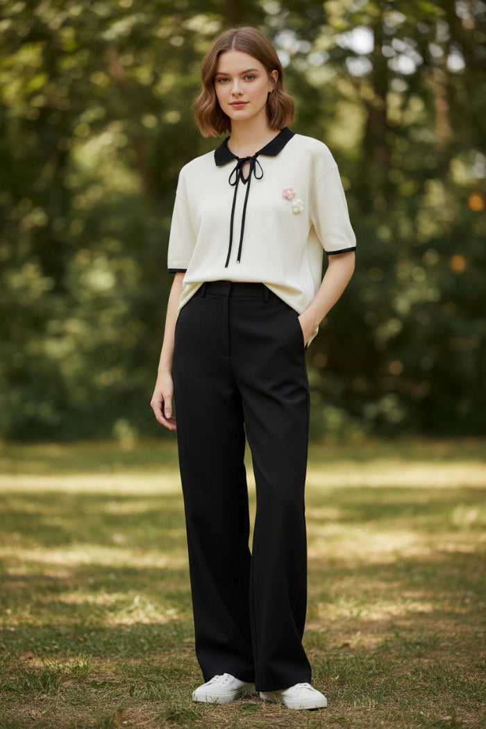An off-white short-sleeve polo shirt with a black collar, black tie detail at the neck, and black piping on the sleeve cuffs. It features a small pink and white floral embroidery on the left chest.