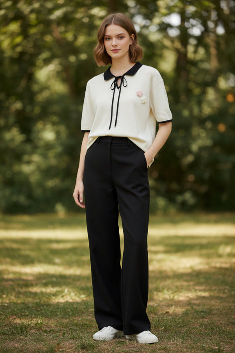An off-white short-sleeve polo shirt with a black collar, black tie detail at the neck, and black piping on the sleeve cuffs. It features a small pink and white floral embroidery on the left chest.