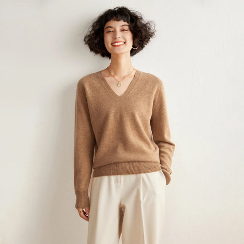 Woman wearing a brown sweater and white pants against a plain background