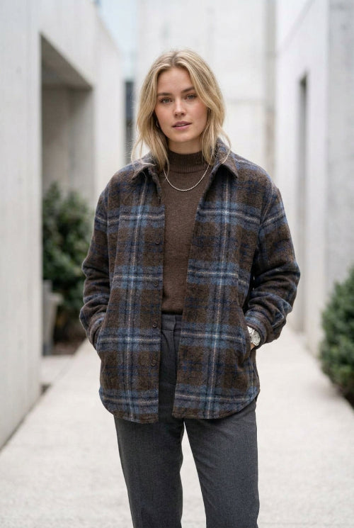 A long-sleeve plaid button-up shirt in dark brown and blue tones, hanging on a black hanger in a retail store. A hand is holding the left sleeve cuff, and a clothing rack and wooden floor are visible in the background.