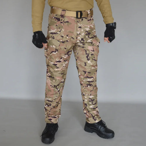 Front view of a model wearing tan, brown, and green camouflage cargo pants with a tan belt, black fingerless gloves, and black lace-up boots against a gray background.⠋