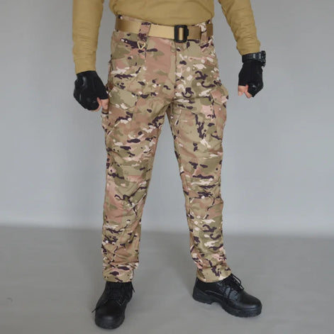 Front view of a model wearing tan, brown, and green camouflage cargo pants with a tan belt, black fingerless gloves, and black lace-up boots against a gray background.⠋