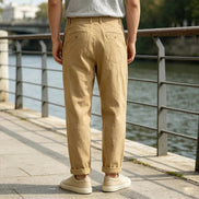 A person standing outdoors by a river, shown from the back wearing light tan straight-leg trousers, a light gray T-shirt, and off-white sneakers.