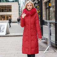 A woman wearing a long red puffer coat with a large fluffy faux-fur collar stands on a city sidewalk. The coat has horizontal quilting and a high collar.