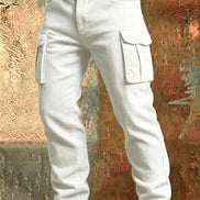 White corduroy cargo pants with flap thigh pockets and a brown belt against a textured rust-colored metal background.